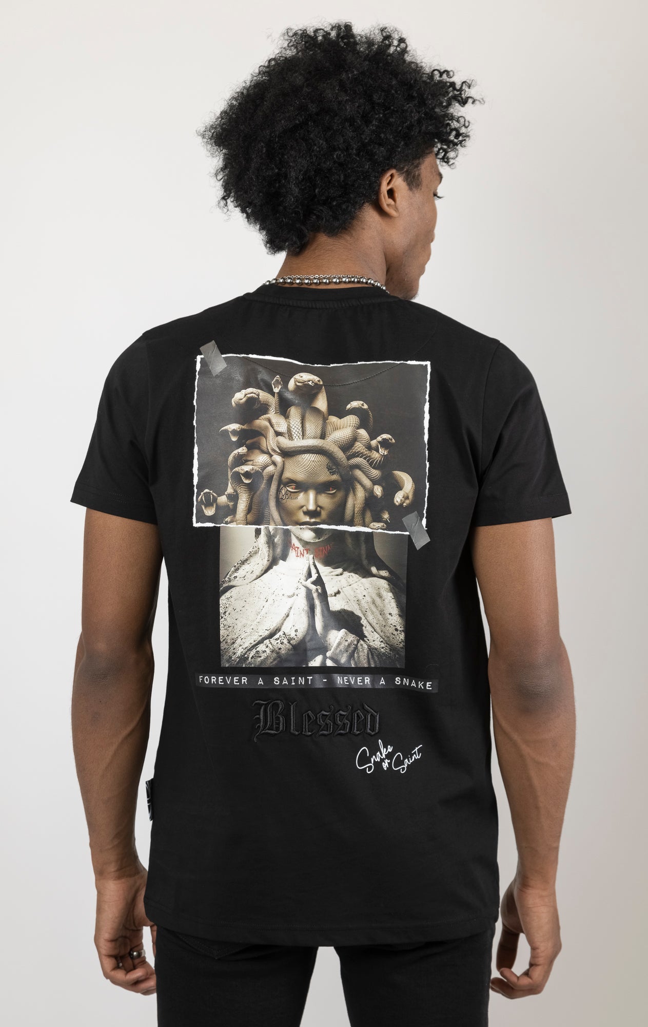 Men's regular fit t-shirt in black. The shirt features a slightly elongated sleeve, a fitted neckline, and a straight body. It has a "Forever a Saint, Never a Snake" digital print on the back with 3D "blessed" embroidery.  Made from 100% cotton.  A "blessed" slogan is printed on the inner neck binding and a woven side seam tab with "blessed" is also included.  The front features a "blessed" with a strikethrough design and the back neck features a signature "blessed" script print.